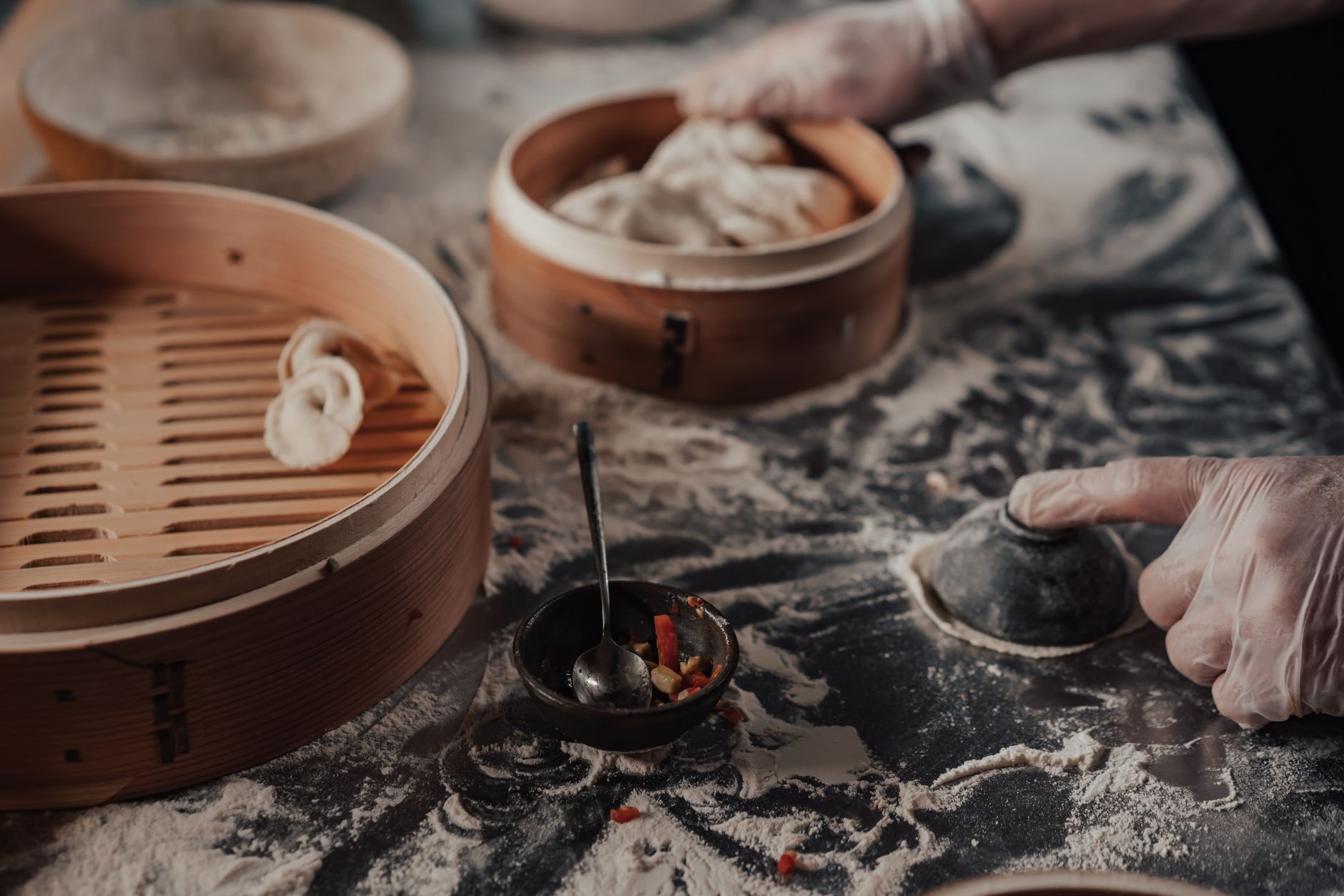 Close-up of hands pleating dumpling dough with bamboo steamers nearby.